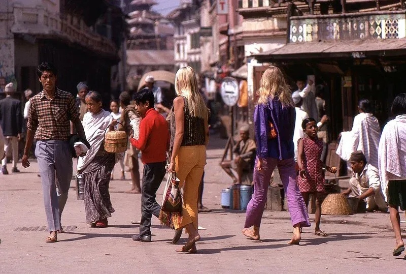 Two blonde hippie women walk through a busy Kathmandu street in the 1970s, with locals and shopfronts around them.